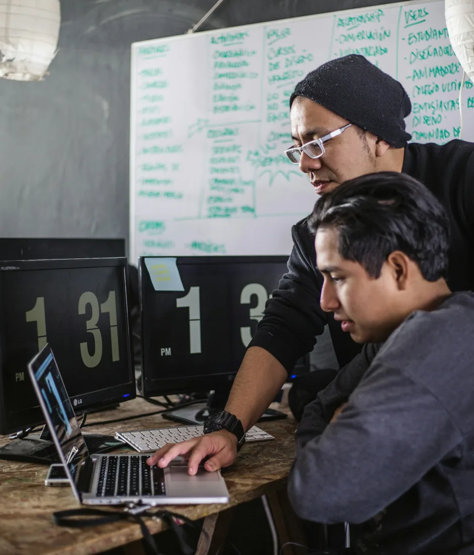 Two developers working through a problem at a laptop, in front of a whiteboard of notes